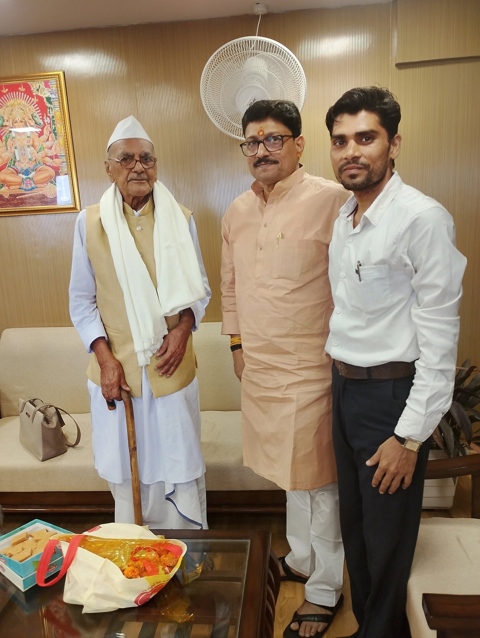 Three men standing together in an office with sofas, an elderly man with a walking stick on the left