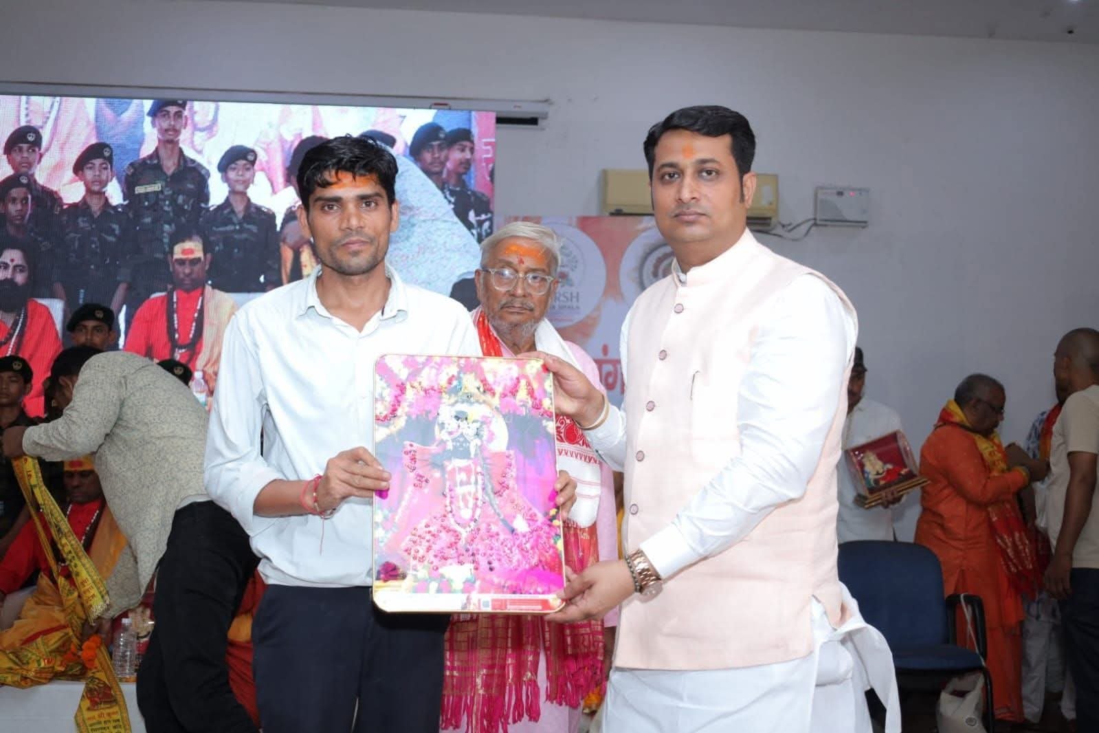 Two men jointly holding a framed picture on stage at an indoor gathering, seated attendees behind