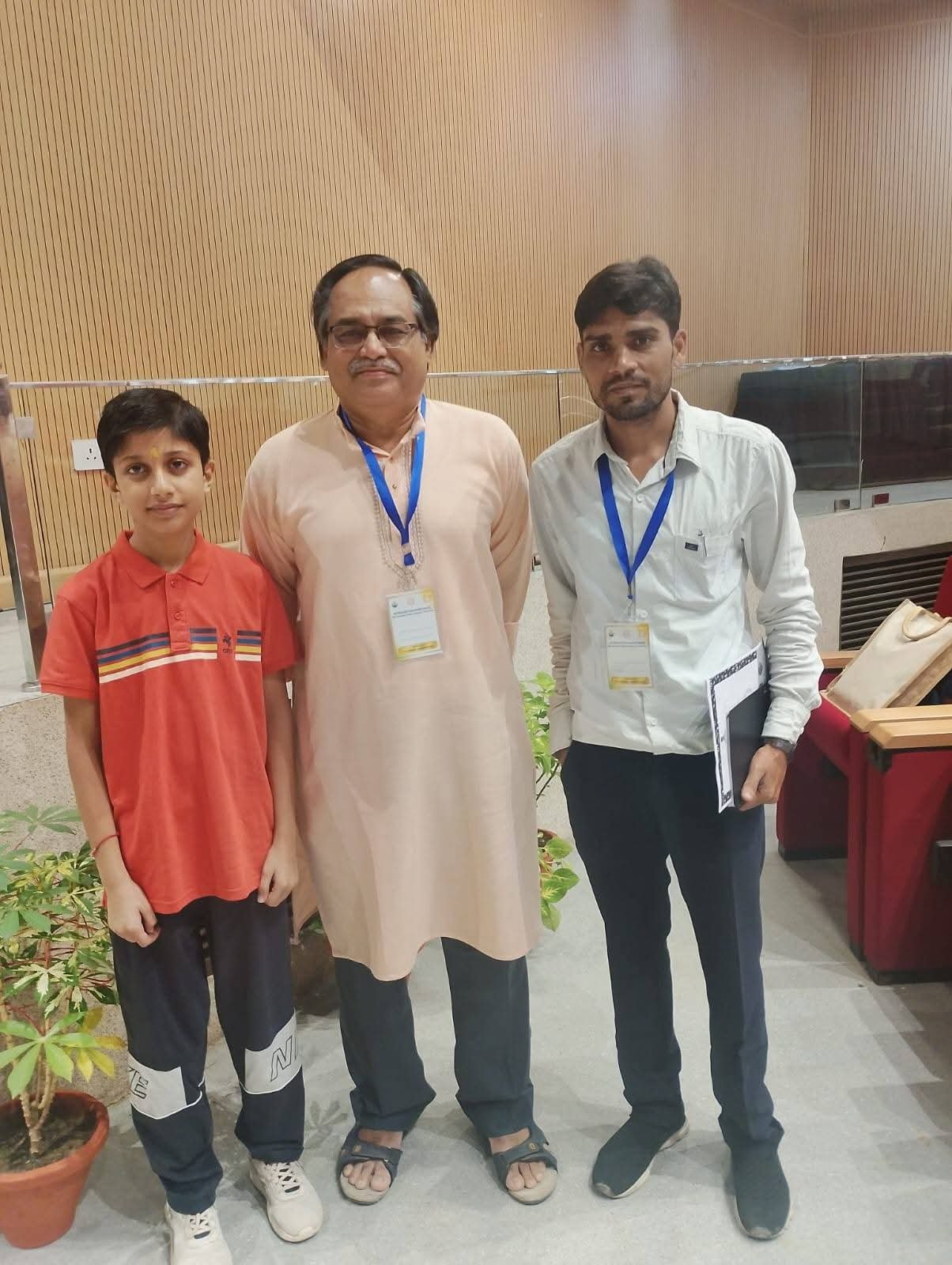 A man in a peach kurta standing between a young boy and another man, all wearing lanyards, at an indoor conference venue