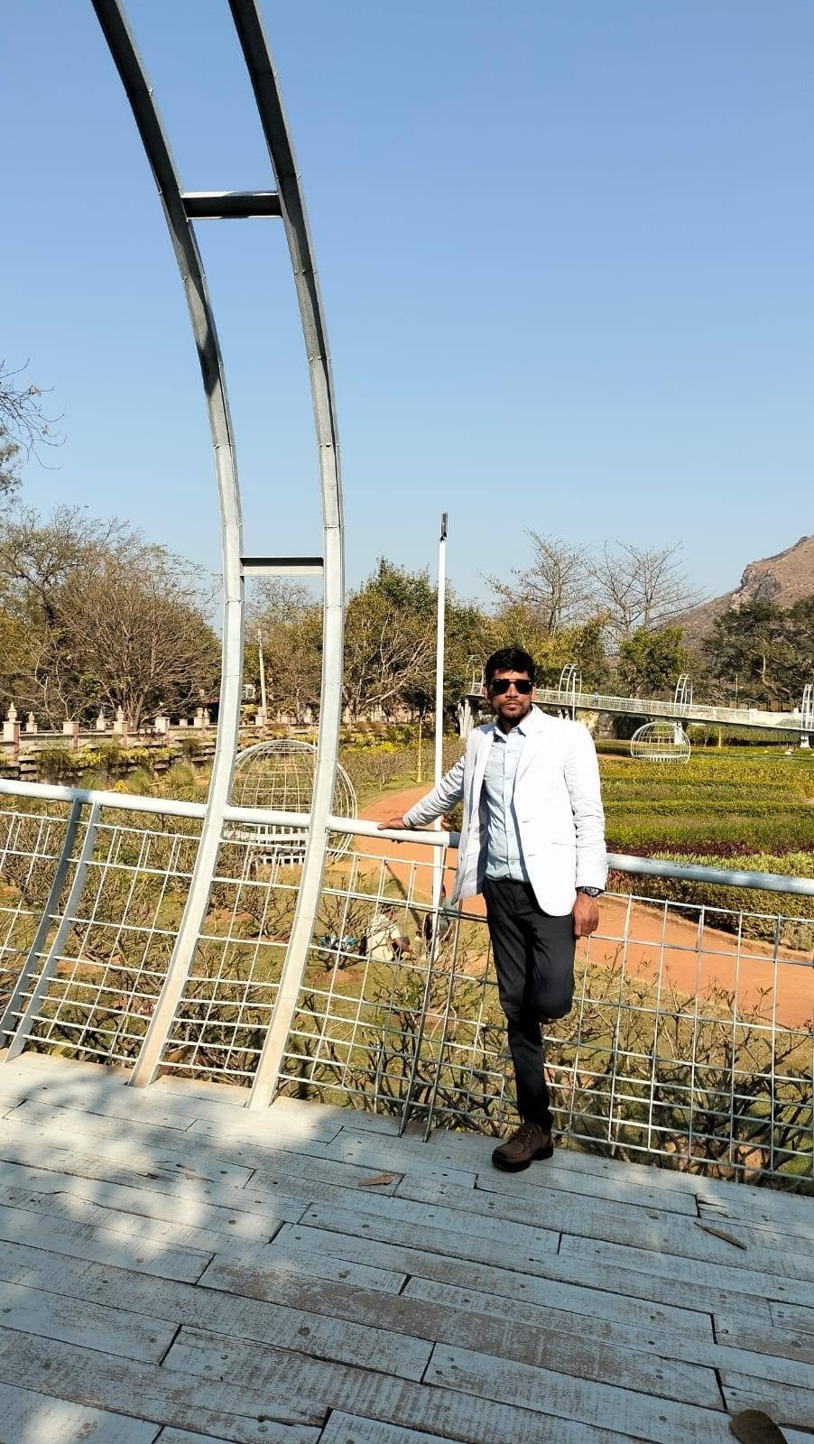 A man in a white blazer leaning on a railing at a landscaped park with hills in the distance